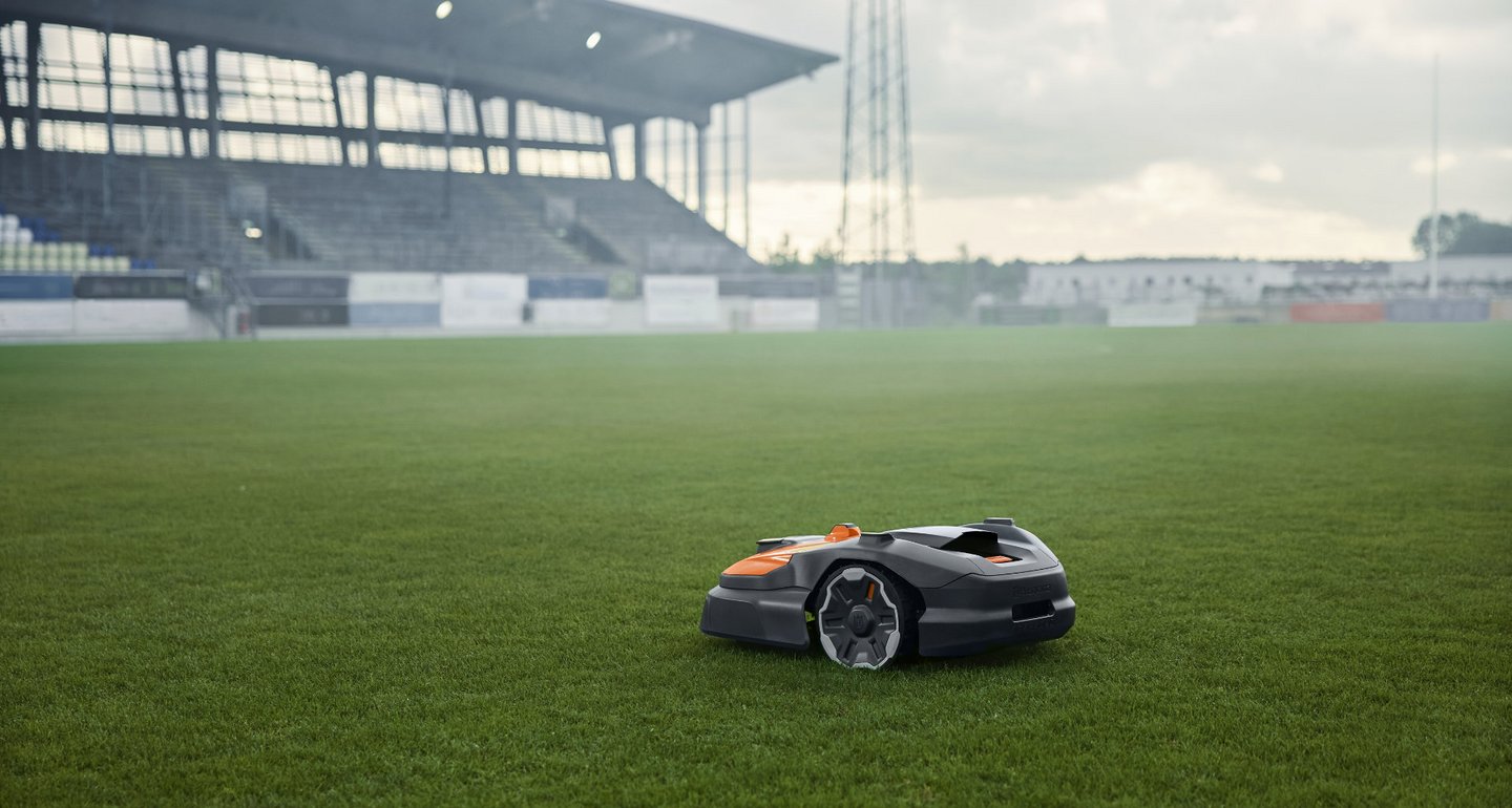 Ein robotischer Rasenmäher mäht den Rasen in einem großen, leeren Fußballstadion. Im Hintergrund sind die Tribünen und ein beleuchteter Stadionmast zu sehen.