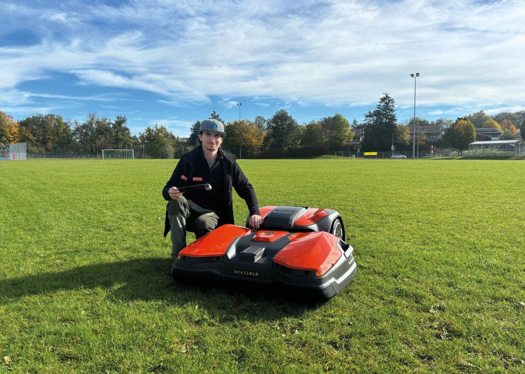 Ein Mann kniet auf einem Fußballfeld neben einem großen, roten, autonomen Rasenmähroboter und hält eine Fernbedienung in der Hand.