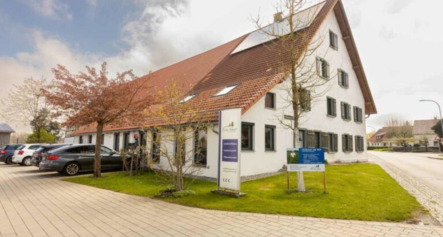 Großes traditionelles Gebäude mit weißer Fassade und rotem Ziegeldach, umgeben von Rasenflächen und einigen Bäumen, neben einem Parkplatz mit mehreren Autos. Vor dem Gebäude stehen Informationsschilder.