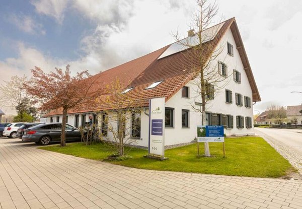 Großes traditionelles Gebäude mit weißer Fassade und rotem Ziegeldach, umgeben von Rasenflächen und einigen Bäumen, neben einem Parkplatz mit mehreren Autos. Vor dem Gebäude stehen Informationsschilder.