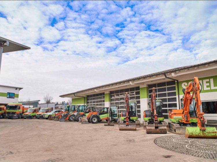 Mehrere Bagger und Baumaschinen sind vor einem großen Gebäude mit grünen Akzenten geparkt, unter einem bewölkten Himmel.