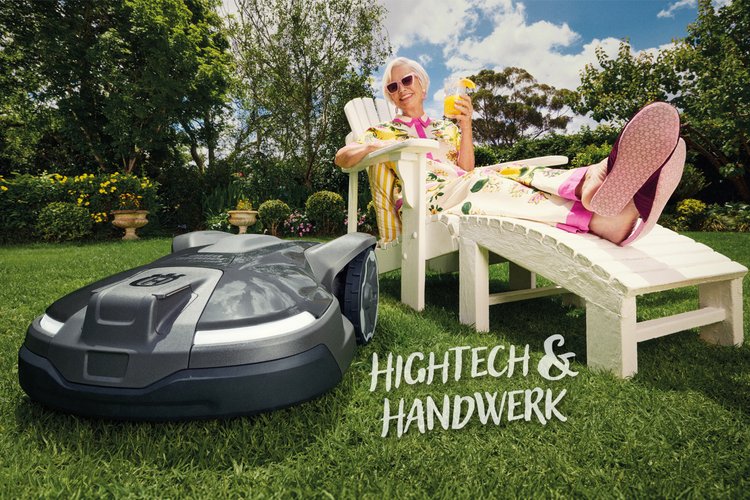 Titelseite des Frühjahrprospekts 2026. Eine ältere Dame liegt auf einem Liegestuhl mit einem Cocktail in der Hand und beugt sich zu einem Mähroboter im Bildvordergrund.