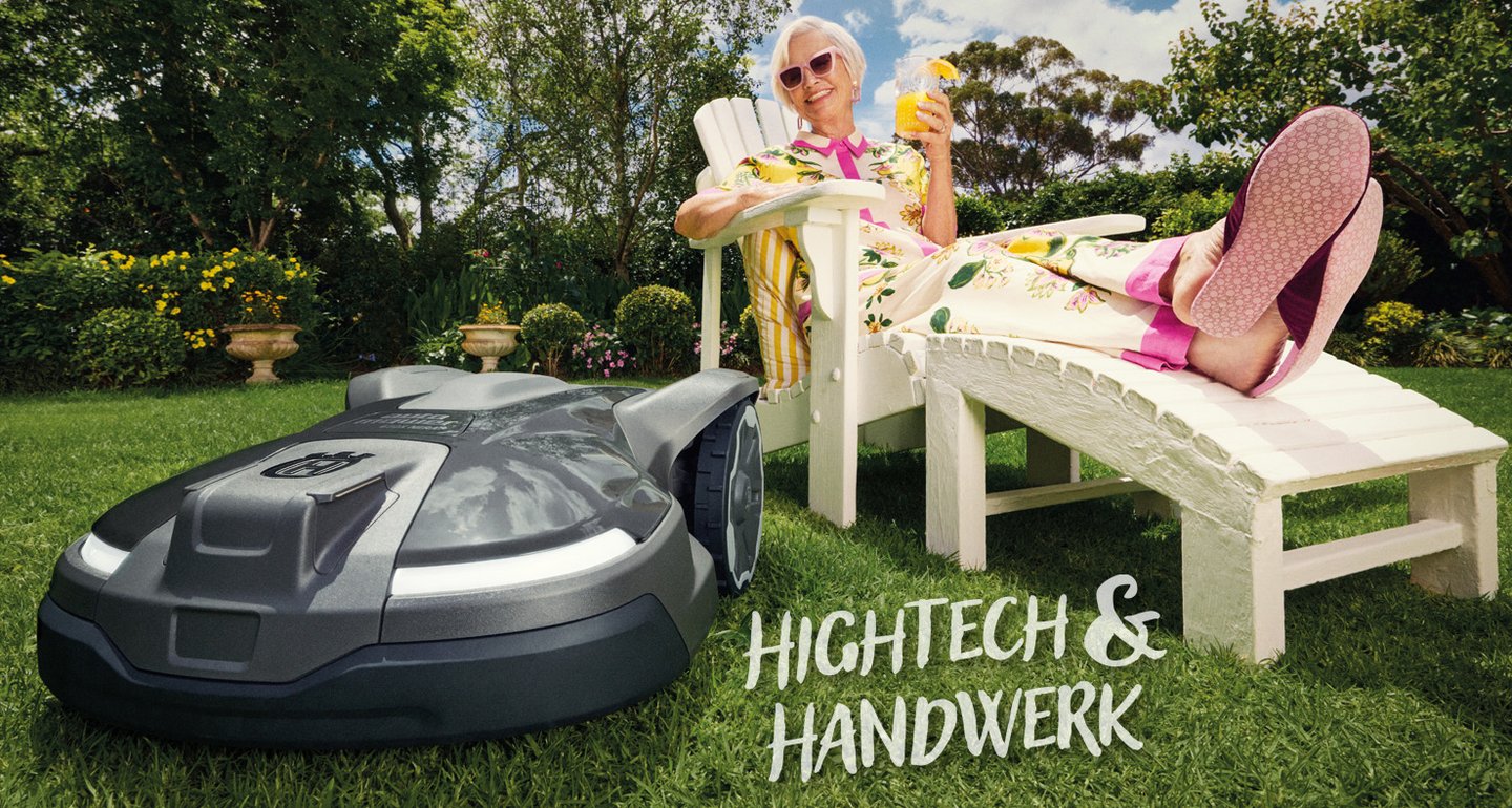 Titelseite des Frühjahrprospekts 2026. Eine ältere Dame liegt auf einem Liegestuhl mit einem Cocktail in der Hand und beugt sich zu einem Mähroboter im Bildvordergrund.