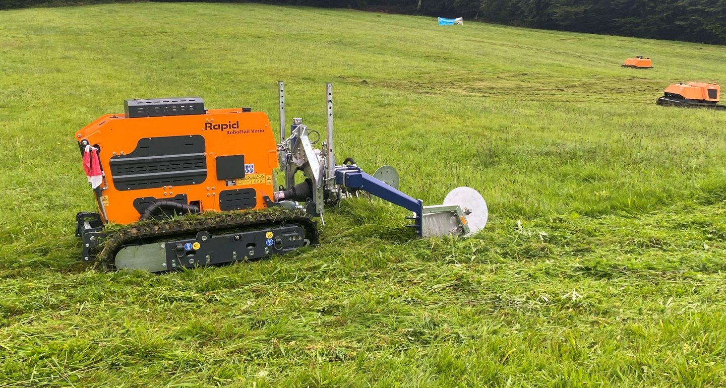 Ein oranger ferngesteuerter Mähroboter mäht eine große Wiese, im Hintergrund sind zwei weitere ähnliche Geräte zu sehen.