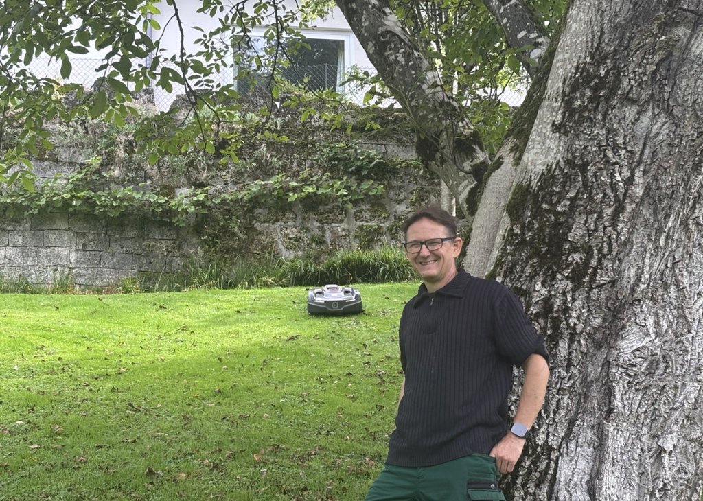 Mann lehnt lächelnd an einem großen Baumstamm im Garten, im Hintergrund fährt ein Mähroboter auf dem Rasen.