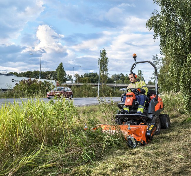 Ein Arbeiter in Schutzkleidung mäht mit einem Aufsitzmäher hohes Gras am Straßenrand an einem bewölkten Tag.