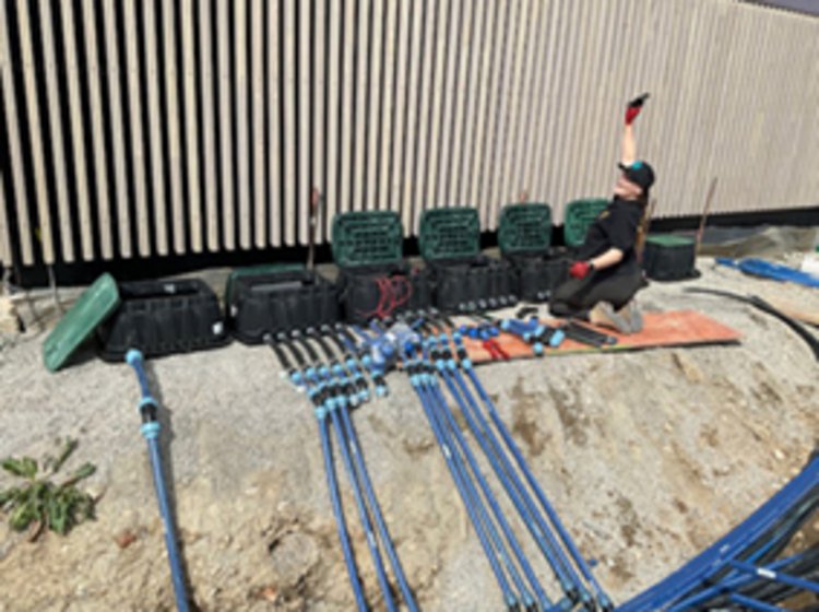 Vor einer Bretterwand sind sieben Container, die auf Sand stehen. Zu den Containern führen viele blaue Rohre. Rechts vor den Containern kniet eine Frau in Arbeitskleidung und winkt.