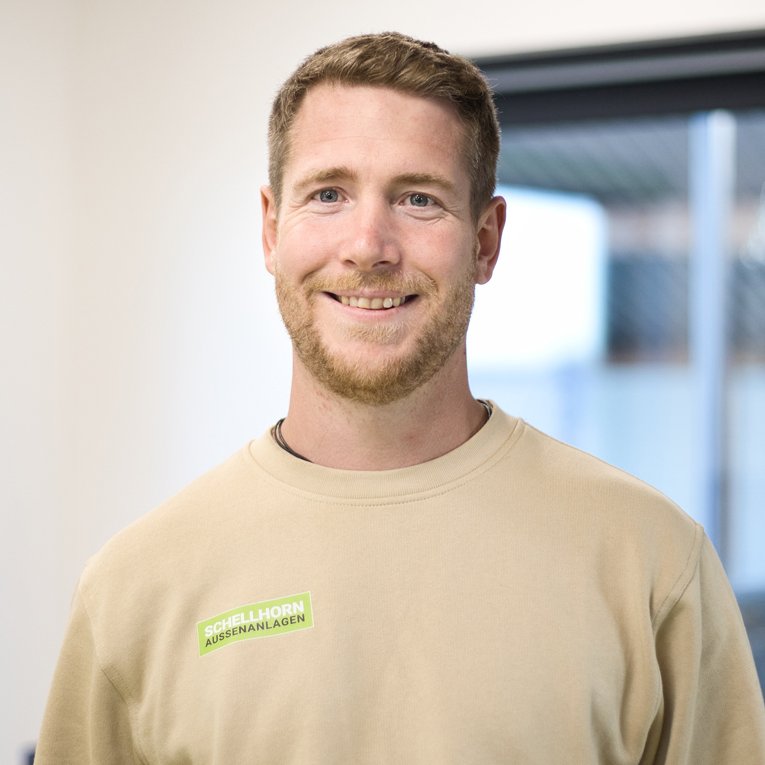 Junger Mann mit kurzem Haar und Bart lächelt freundlich, trägt beigefarbenes Sweatshirt mit grünem Schriftaufdruck.