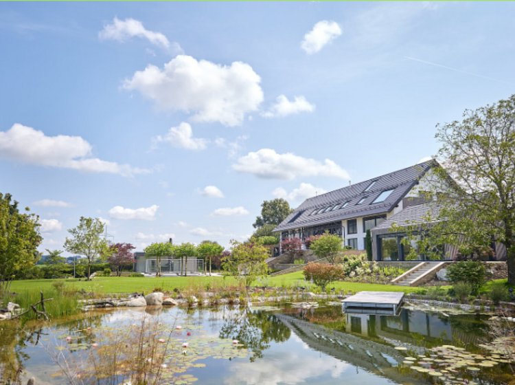 Ein modernes Haus mit großem Garten und Teich, umgeben von Bäumen und Blumen, unter einem blauen Himmel mit weißen Wolken.