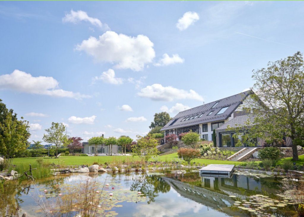 Ein modernes Haus mit großem Garten und Teich, umgeben von Bäumen und Blumen, unter einem blauen Himmel mit weißen Wolken.