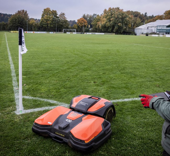 Ein Mitarbeiter steuert einen profi Mähroboter für Fußballplätze am Eckpfosten vom Fußballstadion Bad Waldsee. Im Hintergrund sieht man den Platz mit seinen bekannten Linien.