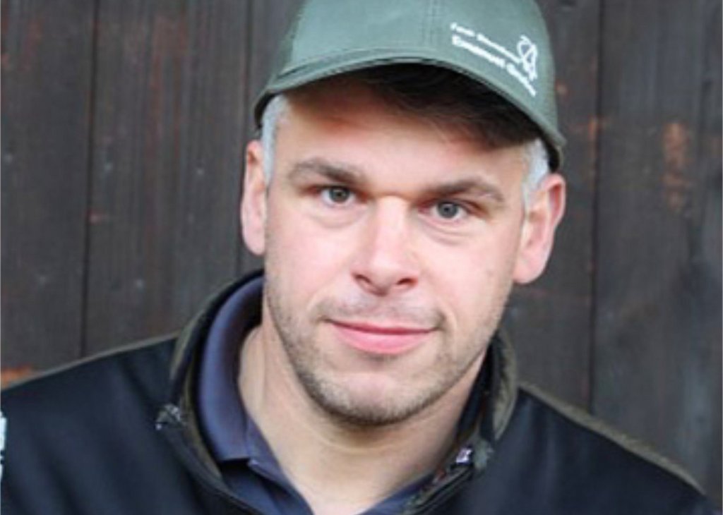 Porträt eines Mannes mit Baseballcap und Outdoorjacke vor dunklem Holz, der direkt in die Kamera lächelt.