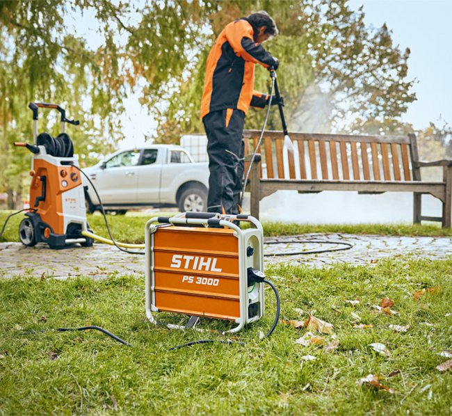 Eine Person reinigt mit einer STIHL Hochdruckreinigungsmaschine eine Parkbank im Freien. Im Vordergrund steht ein STIHL PS 3000 Aggregat, das die Stromversorgung für das Reinigungsgerät bereitstellt.