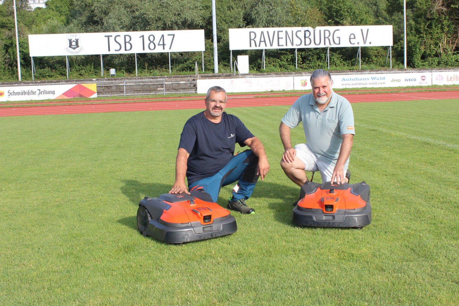 Der Platzwart der Stadt Ravensburg, Markus Reichle, und Ekrem Ozan, Fa. Bentele, knien hinter zwei Mährobotern.