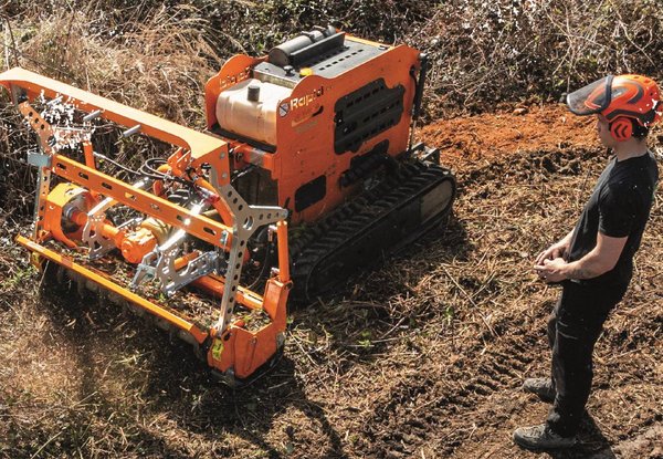Ein Mann steuert eine ferngesteuerte, orangefarbene Mulchmaschine, die auf unebenem, gerodetem Gelände Spuren hinterlässt.