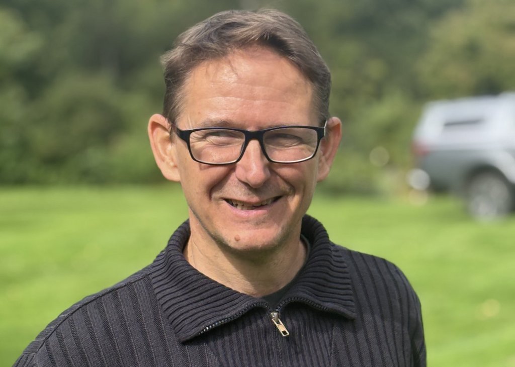 Ein lächelnder Mann mit kurzen braunen Haaren und Brille trägt einen dunklen Pullover. Er steht draußen auf einer grünen Wiese mit Bäumen im Hintergrund.