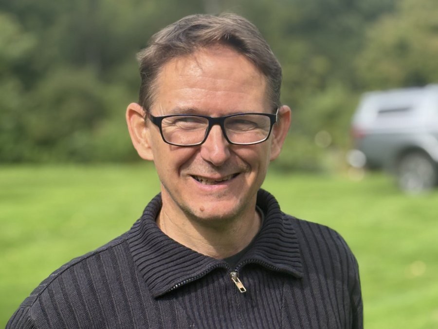 Ein lächelnder Mann mit kurzen braunen Haaren und Brille trägt einen dunklen Pullover. Er steht draußen auf einer grünen Wiese mit Bäumen im Hintergrund.