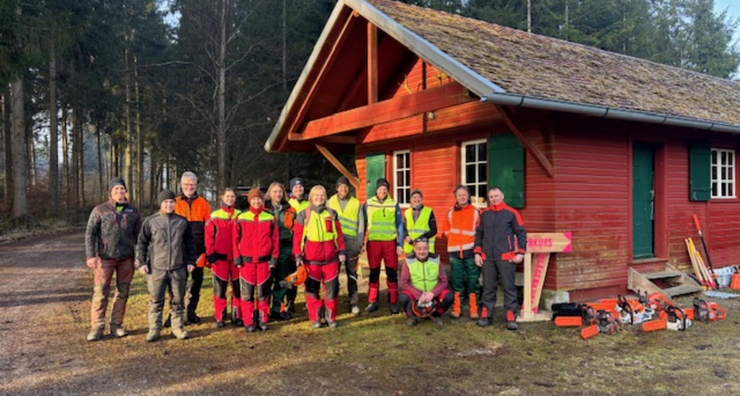 Eine Gruppe von Motorsägen-Kursteilnehmern und die Kursleiter stehen vor einer Waldhütte.