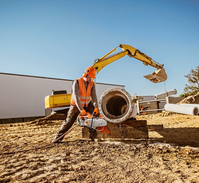 Ein Bauarbeiter mit Schutzhelm und Warnweste schneidet eine Betonrohrleitung mit einer großen Motorsäge auf einer Baustelle, neben einem gelben Bagger und weiteren Betonrohren im Sandboden.
