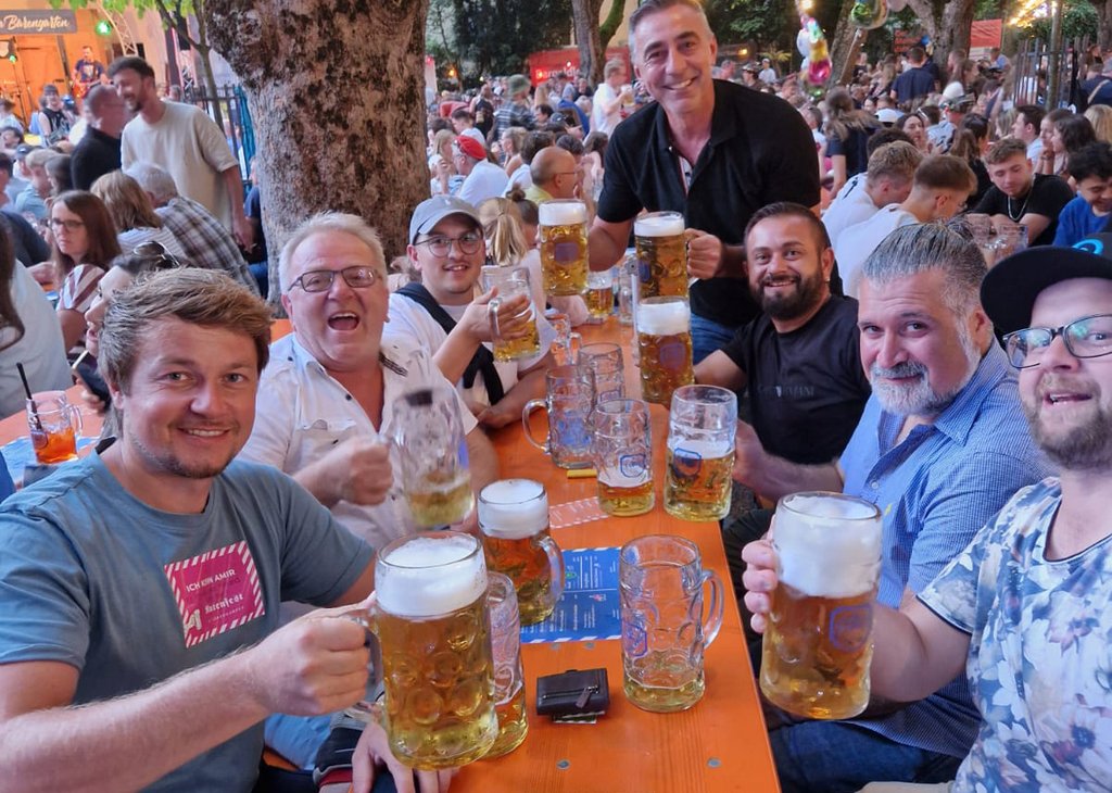 Sechs Männer sitzen an einem langen Holztisch im Freien, heben große Bierkrüge und lachen gemeinsam. Im Hintergrund ist eine belebte Biergartenatmosphäre mit vielen weiteren Gästen und Bäumen zu erkennen.