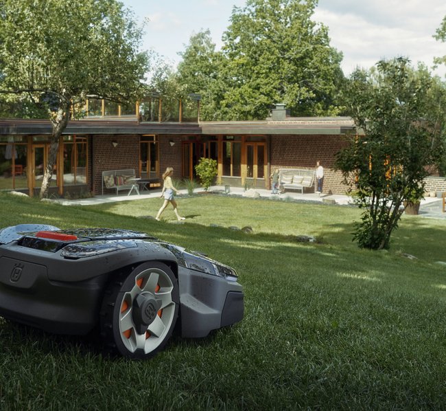 Ein autonomer Mähroboter fährt über einen gepflegten Rasen vor einem modernen Haus mit Terrasse, während eine Person im Hintergrund spazieren geht. Bäume und Sträucher umgeben den Garten.