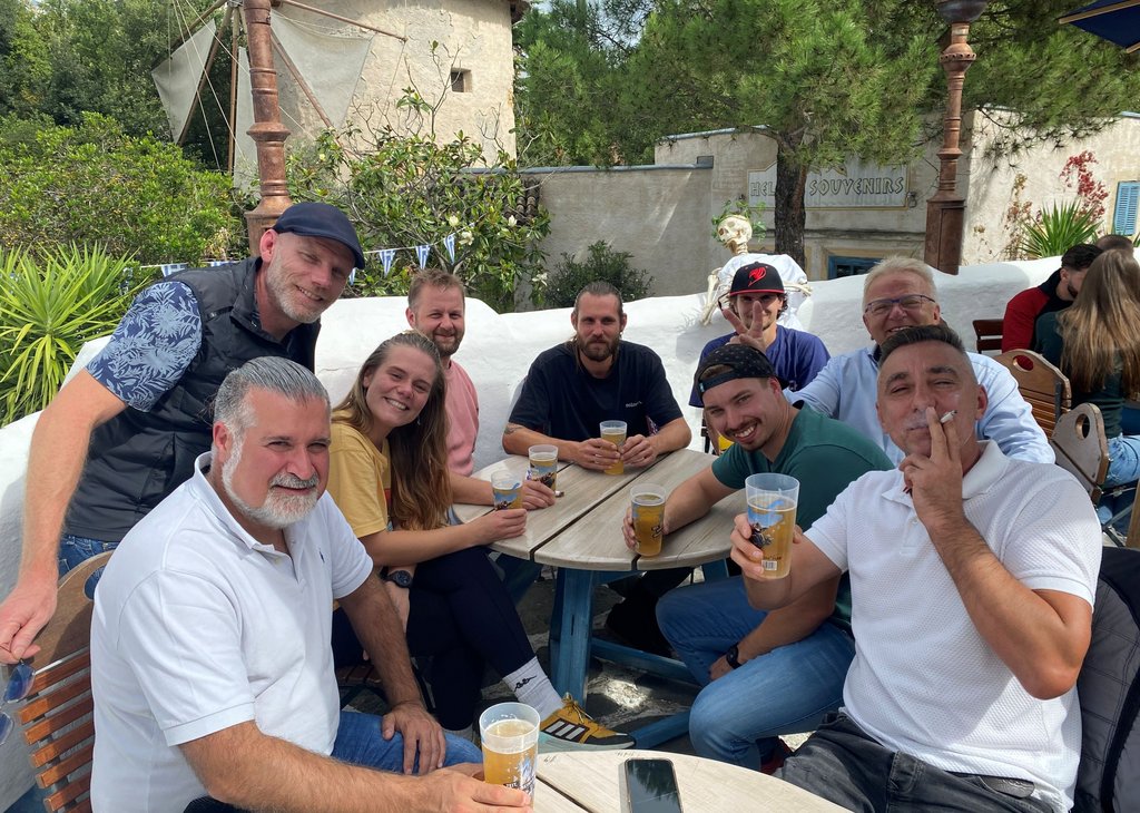 Neun Personen sitzen lachend und entspannt an einem Außentisch, trinken Bier und genießen die gemeinsame Zeit an einem sonnigen Tag.