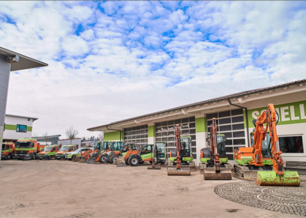 Mehrere Bagger und Baumaschinen sind vor einem großen Gebäude mit grünen Akzenten geparkt, unter einem bewölkten Himmel.