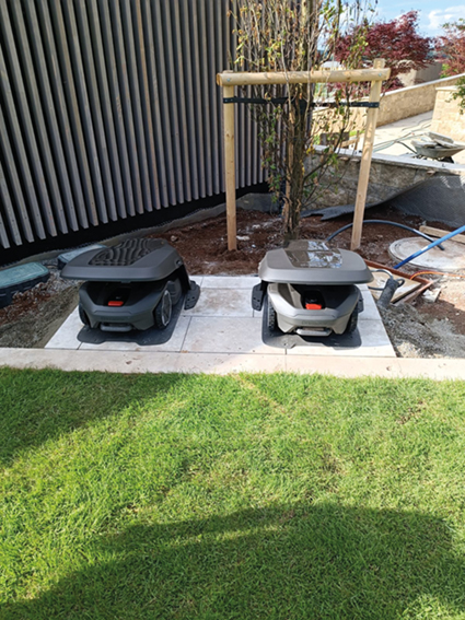 Zwei Mähroboter stehen in ihren Garagen in einer Ecke eines Grundstücks vor einem Baum.